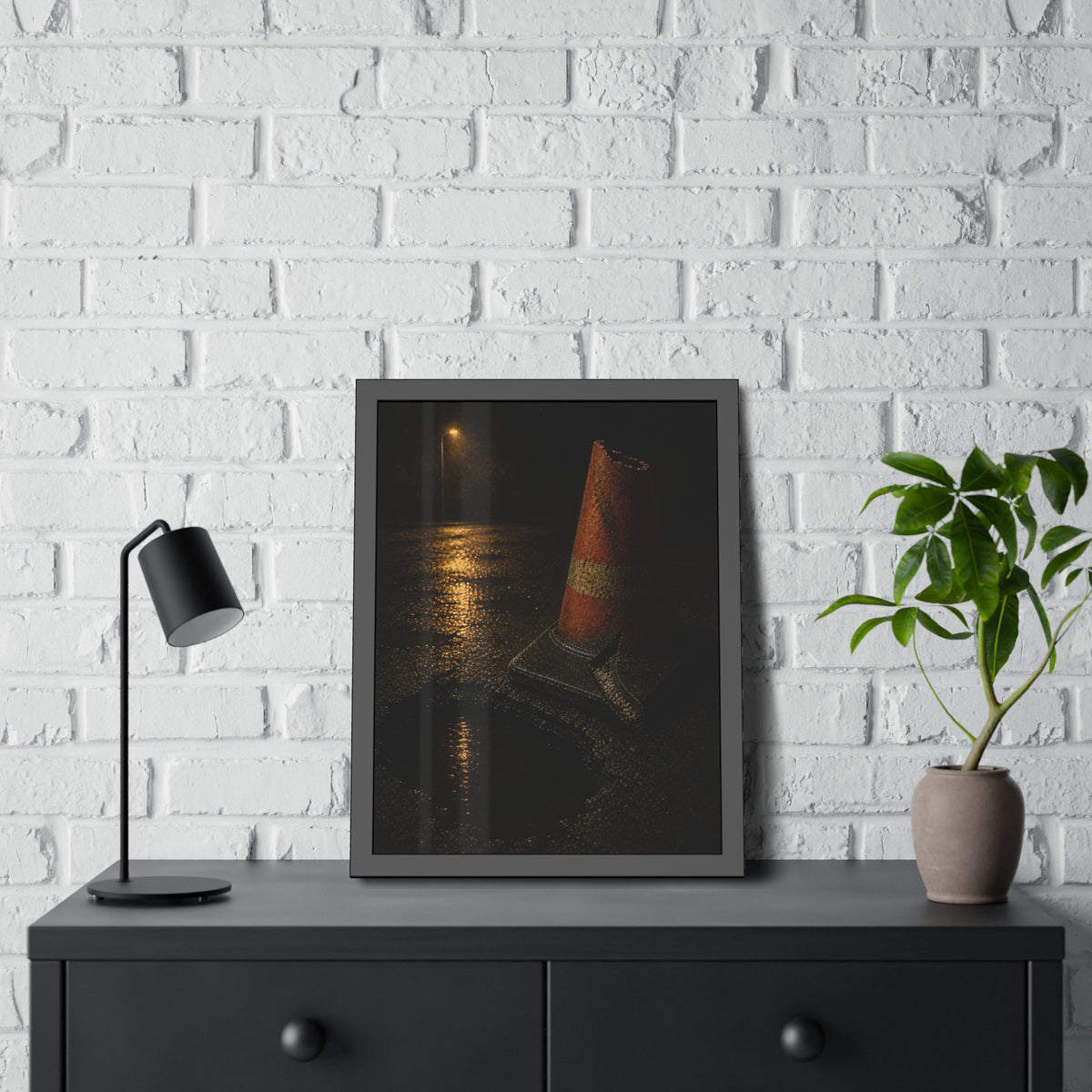 A traffic cone beside a water filled pothole reflecting a street lamp. The Sentinel of the Pothole by Garabaggio, framed fine art print hanging in a room.