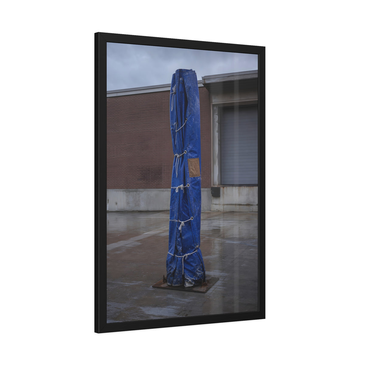 A tall blue tarp bundle roped upright on a wet lot by a loading dock. Folded Standard at the Loading Bay by Garabaggio, framed fine art print hanging in a room.