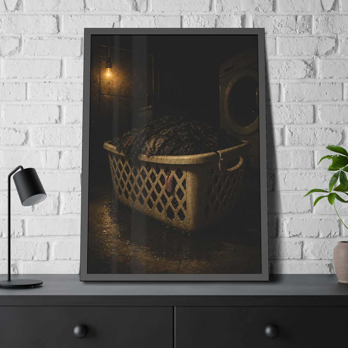 A laundry basket heaped with wet dark clothes under a bare bulb. The Burden of Wet Clothes by Garabaggio, framed fine art print hanging in a room.