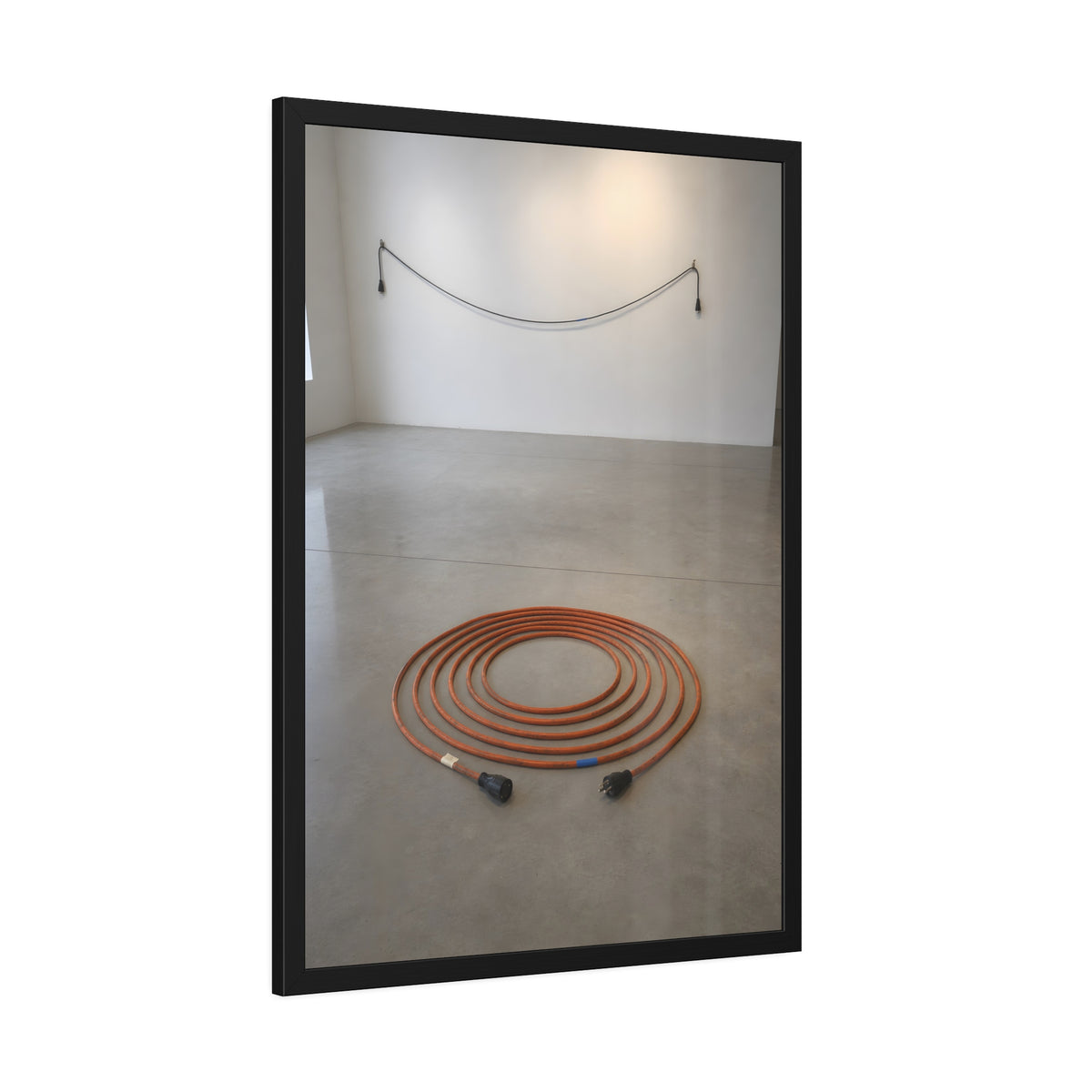 An orange extension cord coiled in a spiral on a bare gallery floor. The Floor Coil by Garabaggio, framed fine art print hanging in a room.