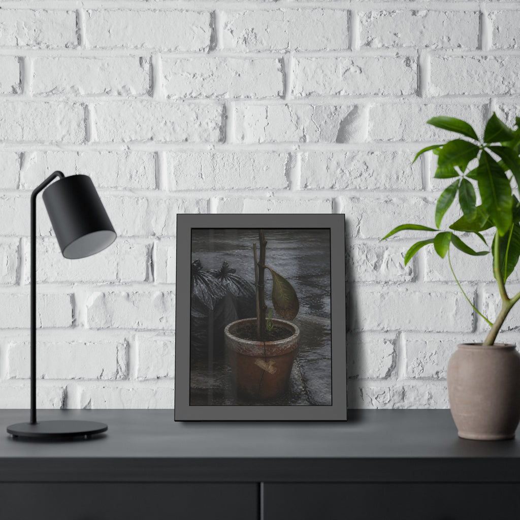 A taped clay pot with one leaf and a green shoot on wet pavement. The Green at the Base by Garabaggio, framed fine art print hanging in a room.