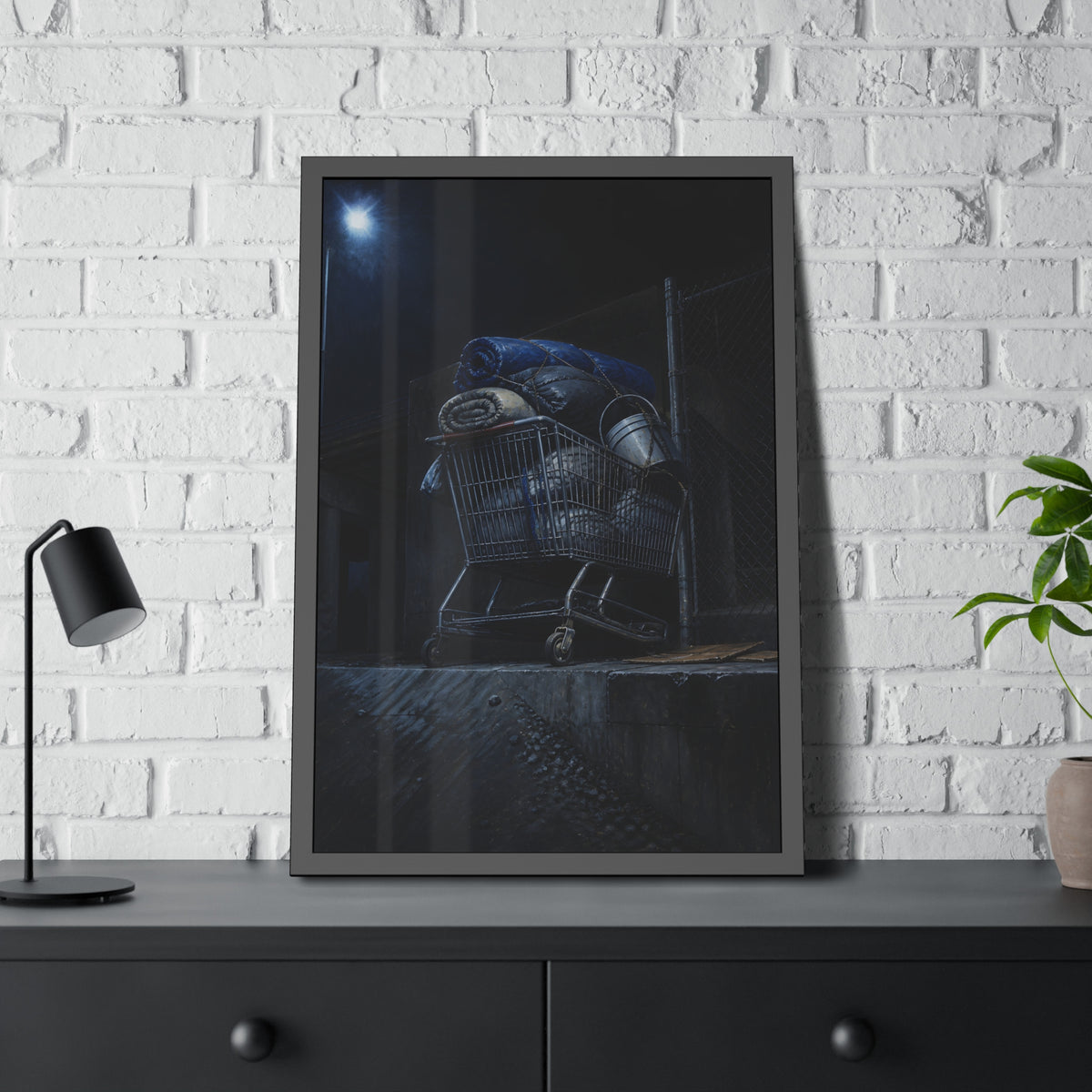 A shopping cart piled with bedding and buckets under a streetlamp. The Loading of Another Life by Garabaggio, framed fine art print hanging in a room.