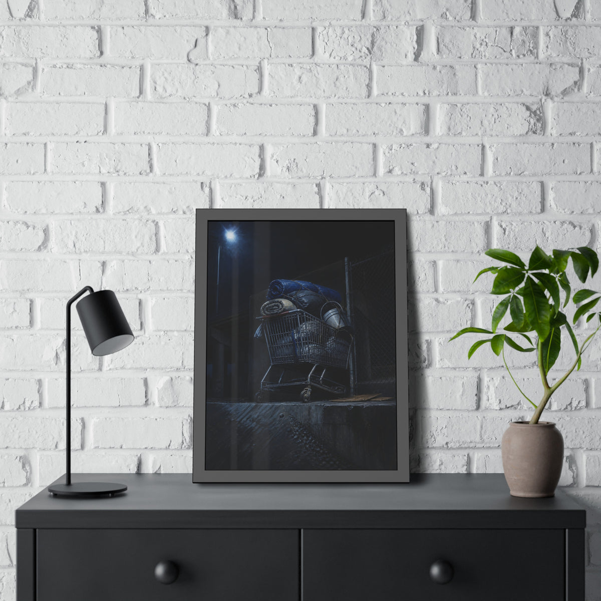 A shopping cart piled with bedding and buckets under a streetlamp. The Loading of Another Life by Garabaggio, framed fine art print hanging in a room.