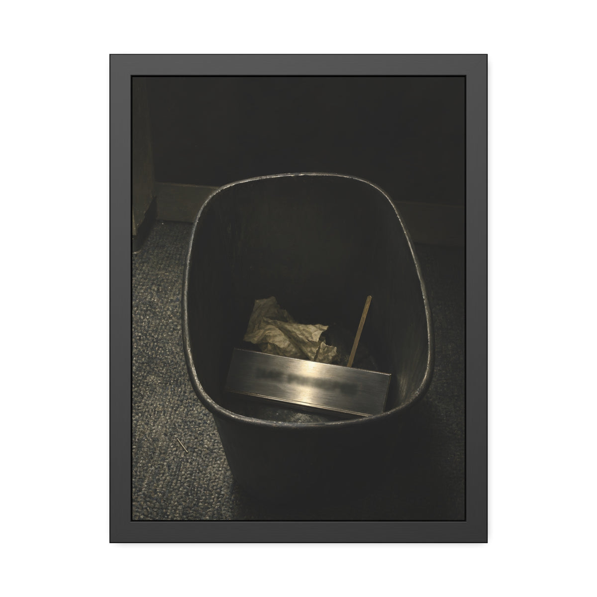 A metal nameplate lying in a black office waste bin on grey carpet. The Nameplate in the Waste Bin by Garabaggio, framed fine art print, 18″ x 24″.