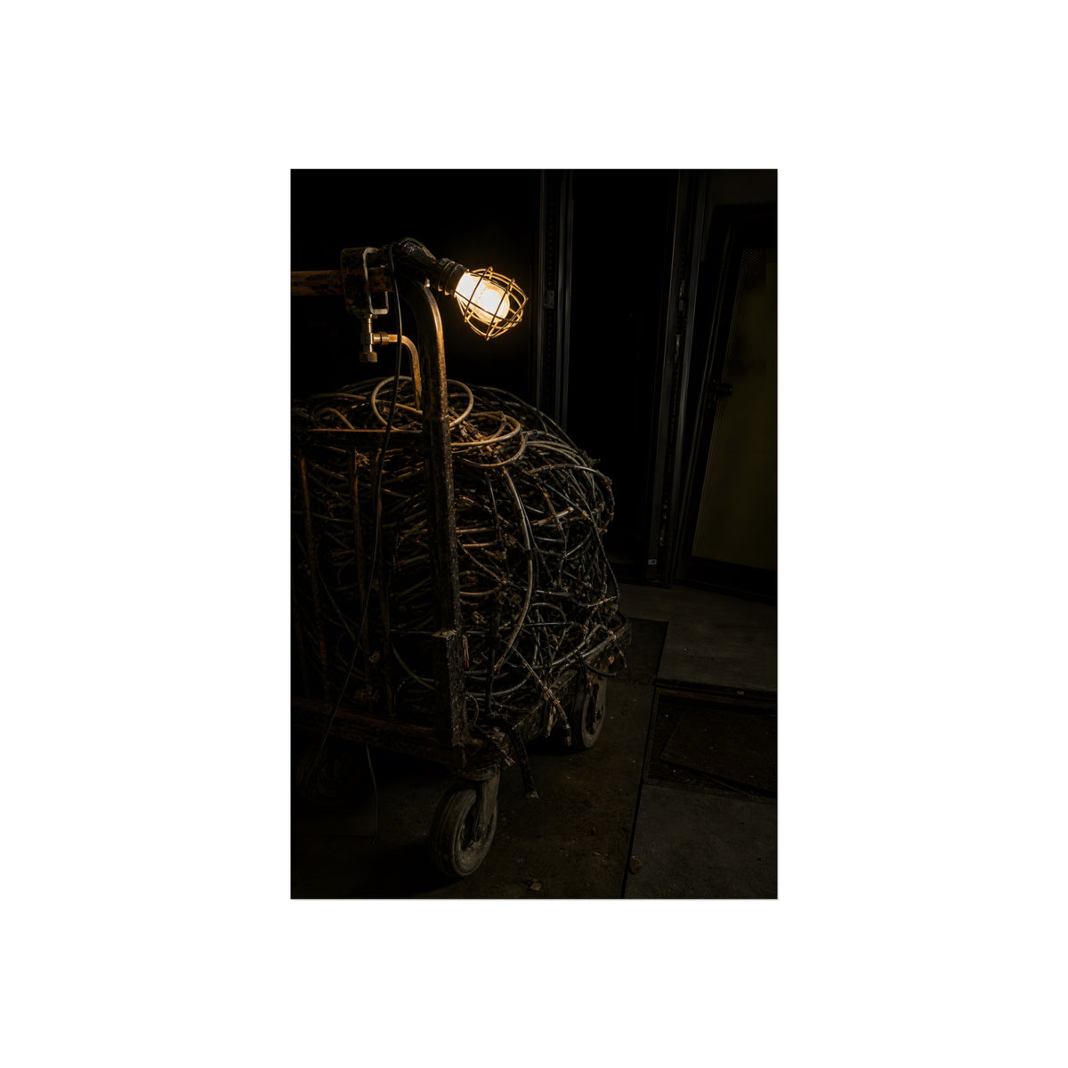 A tangled ball of cables on a hand cart under a caged work lamp. The Cable Harvest by Garabaggio, fine art print, 24″ x 36″.