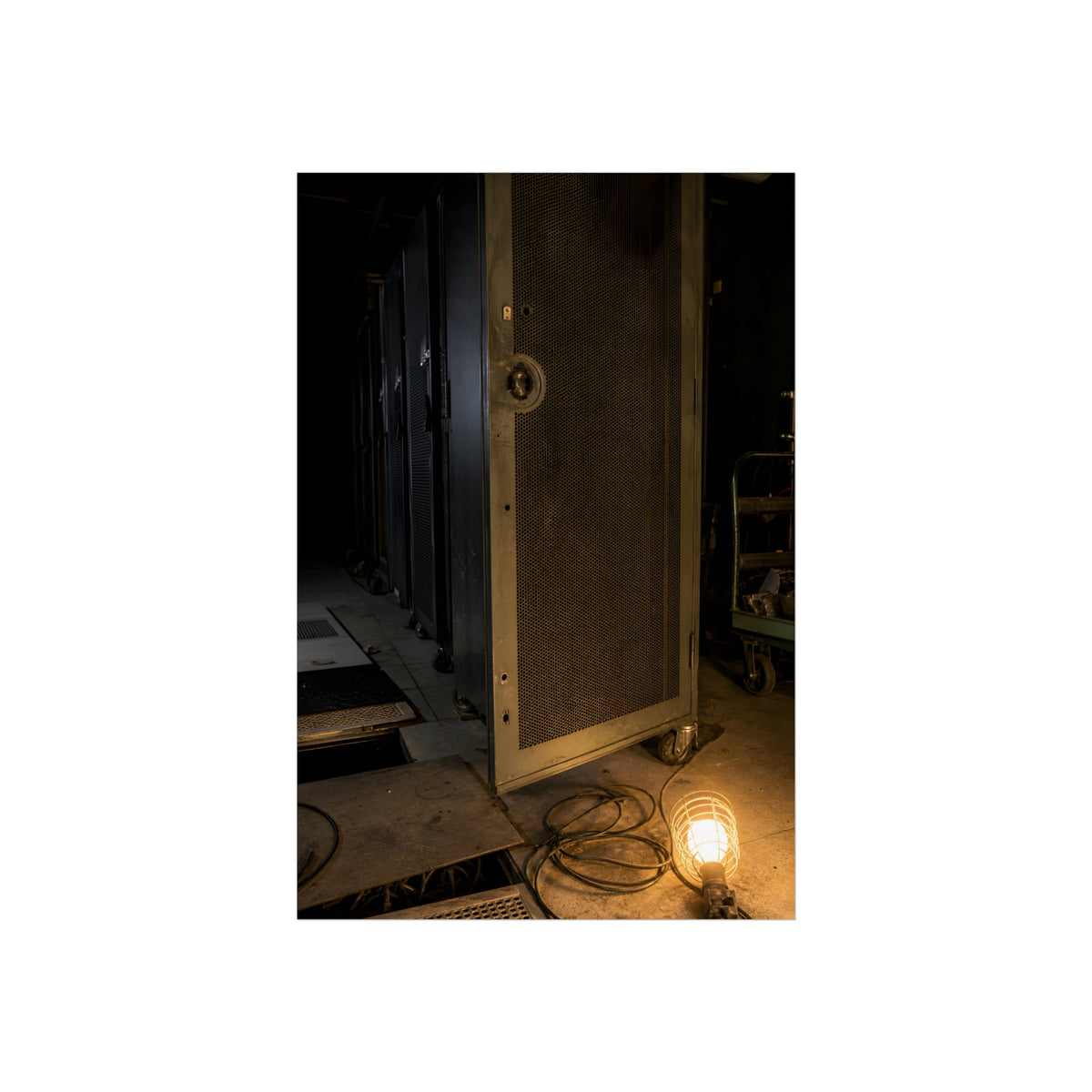 A perforated server rack door in a dark aisle lit by a floor work lamp. The Cold Aisle by Garabaggio, fine art print, 24″ x 36″.