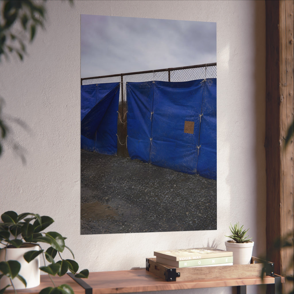 Blue tarps lashed to a chain link fence above gravel under grey sky. Drapery on the Fence by Garabaggio, fine art print hanging in a room.