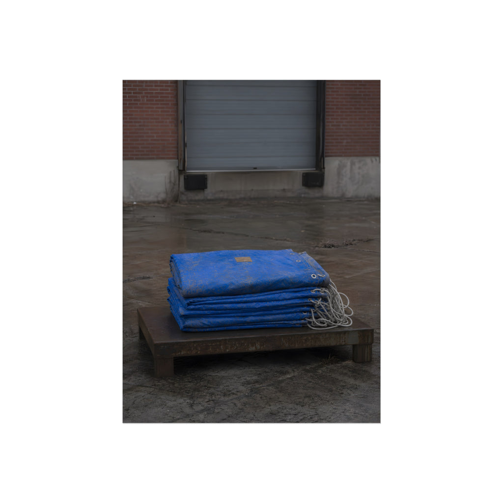 A folded blue tarp stacked on a low wooden platform by a loading dock. The Repaired Fold by Garabaggio, fine art print, 12″ x 16″.