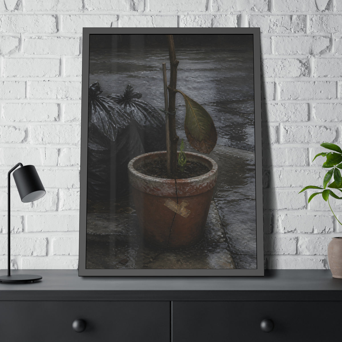 A taped clay pot with one leaf and a green shoot on wet pavement. The Green at the Base by Garabaggio, framed fine art print hanging in a room.