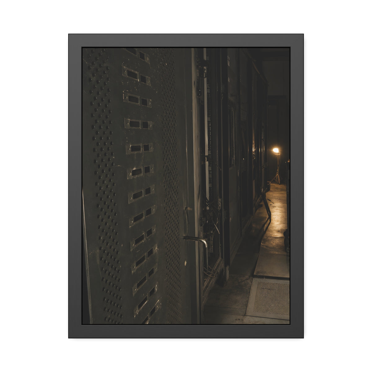 Rows of dark server cabinets with one work lamp glowing far down the aisle. Racks Without Light by Garabaggio, framed fine art print, 18″ x 24″.
